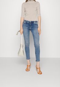 Suéter de cuello alto gris claro, jeans ajustados azules, sandalias de tacón alto naranjas y un bolso estructurado blanco en la mano.