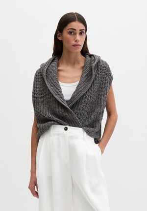 Femme portant un haut blanc, un pantalon blanc ample et un cardigan sans manches en maille épaisse grise avec un col drapé.