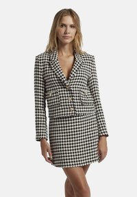 Op maat gemaakte zwart-wit houndstooth blazer met gouden knopen en bijpassende mini-jurk. Voorzien van een notch lapel en twee voorzakken.