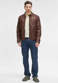 Giacca di pelle marrone con zip e tasche laterali, indossata sopra una maglietta crema chiara, abbinata a jeans blu e sneakers verdi.