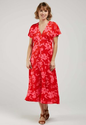 Femme debout portant une robe midi rouge à imprimé floral rose, manches courtes, col en V, avec des sandales à lanières marron sur un fond clair.