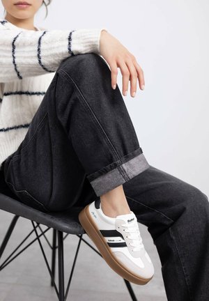 Zapatilla blanca con rayas negras y suela marrón clara. Llevada con pantalones grises arremangados en los tobillos y un suéter blanco texturizado con rayas oscuras.