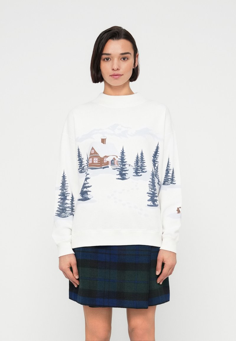Νεαρή γυναίκα που φορά λευκό φουτεράκι με εκτύπωση χειμερινής καλύβας και πεύκων, και σκουρόχρωμη καρό φούστα σε μπλε και πράσινο.