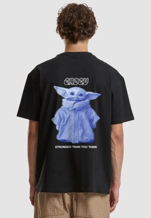 Personne portant un t-shirt noir avec un motif Grogu bleu et le texte « Stronger than you think » dans le dos, debout devant un fond uni.