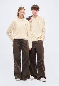 Deux mannequins sont côte à côte, portant des pulls crème, des pantalons larges marron et des baskets blanches sur un fond blanc uni.