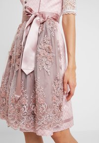 Robe rose avec un tissu à motifs et des manches en dentelle. Le superposé présente des broderies florales et des perles, maintenue par un ruban en satin à la taille.