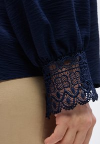 Χέρι που φοράει μπλούζα ναυτικού χρώματος με υφή υφάσματος και λεπτομερή δαντέλα στο μανίκι, ακουμπισμένο σε μπεζ φόντο υφάσματος.