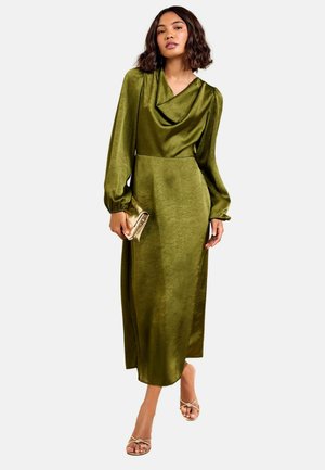 Frau trägt ein olivgrünes Satinkleid mit langen Ärmeln und hält eine goldene Clutch, kombiniert mit goldenen Sandalen mit offenen Zehen, stehend vor weißem Hintergrund.