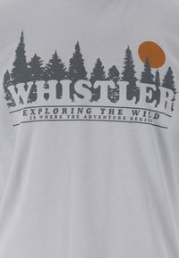 Whistler-Text vor der Silhouette dunkler Kiefern mit einer orangefarbenen Sonne und dem Slogan "Das Abenteuer beginnt, wenn man die Wildnis erkundet" auf grauem Stoff.