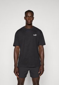 Sort bomulds-t-shirt med korte ærmer, med et hvidt Puma-logo på venstre bryst, parret med sorte shorts. Simple design og tekstur.