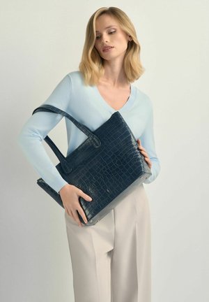 Kobieta ubrana w jasnoniebieski sweter i beżowe spodnie trzyma dużą ciemnoniebieską torbę typu tote o fakturze przypominającej skórę krokodyla na jednolitym tle.