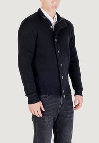 Uomo con un cardigan nero a bottoni sopra una camicia bianca, abbinato a jeans scuri sbiaditi, in piedi contro uno sfondo chiaro e semplice.