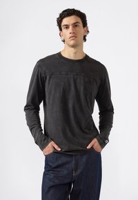 Ein Mann trägt ein verblasstes schwarzes Langarmshirt und dunkelblaue Jeans und steht mit den Händen in den Taschen vor einem schlichten weißen Hintergrund.