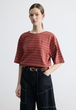 Frau mit heller Haut und zurückgebundenem Haar, die ein rotes, gestreiftes Kurzarmhemd trägt, das in eine dunkle, weit geschnittene Hose mit braunem Gürtel gesteckt ist.