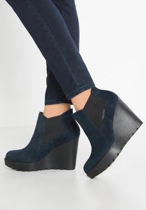 Mørkeblå mokkasinkile kilestøvler båret med mørke skinny jeans, med elastiske sidepaneler og sorte platformsåler.