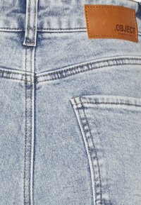 Světle modré džíny z denimu s koženou nášivkou s nápisem "OBJECT" na pase. Mají klasické švy a detail na zadní kapse.
