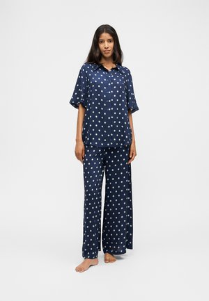 Donna in piedi a piedi nudi che indossa una maglietta blu navy a maniche corte con bottoni e pantaloni ampi con pois bianchi su sfondo semplice.