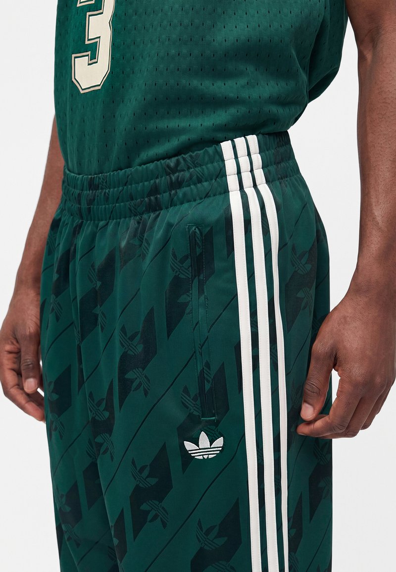 Nahaufnahme einer Person, die dunkelgrüne Adidas-Sportshorts mit weißen drei Streifen und einem Trefoil-Logo sowie ein passendes grünes Trikot mit der Nummer 3 trägt.