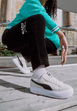 Personne portant un pantalon noir KANI, une veste bleu sarcelle, des baskets blanches Puma et des chaussettes blanches, accroupie sur une surface en bois extérieure près d'un mur en briques.