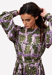 Vrouw die haar donkerbruin haar aanpast, gekleed in een lang jurk met groene, paarse en beige bloem- en luipaardmotieven.