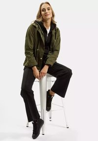 Zielona oliwkowa kurtka na zamek z kieszeniami, noszona na czarnej bluzie z kapturem. Czarne spodnie o szerokich nogawkach i czarne buty sznurowane.