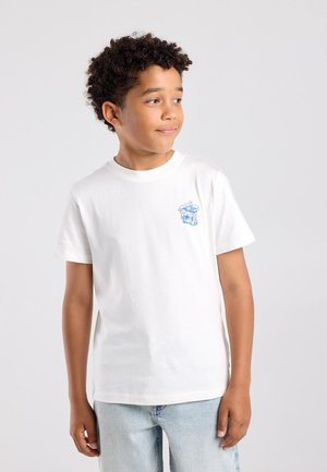 Jongen met krullend haar, die een wit T-shirt met een kleine blauwe afbeelding en lichtblauwe spijkerbroek draagt, kijkt opzij tegen een effen achtergrond.