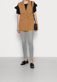Braunes, doppelt geknöpftes Weste mit Schulterakzenten, schwarzes kurzärmliges Oberteil, graue enge Jeans und schwarze Loafers mit Schleifen-Detail.