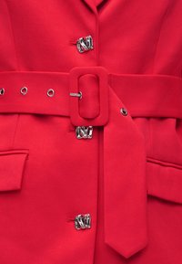 Κόκκινο παλτό ραμμένο στα μέτρα με ζώνη από το ίδιο ύφασμα που διαθέτει ορθογώνια αγκράφα και ασημί στριφτούς γάντζους μπροστά.