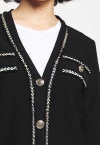 Černý kardigan s texturovanou tkaninou, barevným lemem podél okrajů, knoflíky ve zlatém tónu a dvěma kapsami na hrudi s dekorativními akcenty.