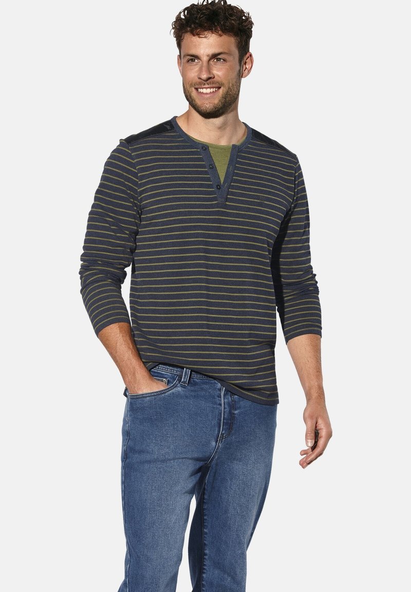 Lächelnder Mann in einem dunkelblauen und grünen gestreiften Langarm-Henley-Shirt, mit blauen Jeans, eine Hand in der Tasche, vor einem weißen Hintergrund.