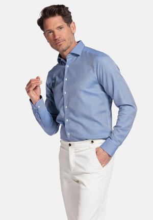 Lichtblauw overhemd met lange mouwen en een gestructureerd patroon, tot aan de kraag dichtgeknoopt. Gepaard met witte broek, weergegeven op een neutrale achtergrond.