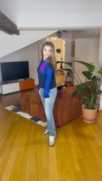 Kék és fekete pulóver virágmintával, világoskék farmernadrág és ezüst sportcipők. Beltéri környezet, fa padlóval és barna kanapéval.