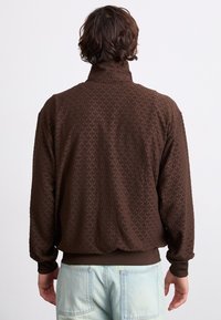 Ruskea teksturoitu takki korkealla kauluksessa, jossa on hienovarainen lehväkuvio. Takissa on joustavat hihat ja rento istuvuus vyötäröllä.