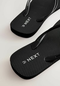 Zwarte flip-flops met een geprofileerde zool, voorzien van zwarte banden en een wit "NEXT" logo. Het ontwerp is eenvoudig zonder extra patronen.