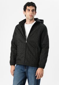 Chaqueta con capucha negra que presenta un patrón texturizado, cierre de cremallera, puños elásticos y bolsillos laterales. Llevada sobre una camiseta blanca y jeans azules.