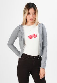 Siva pletena dukserica sa zipom i detaljima od pletenice, preko kremaste košulje s grafikom crvene trešnje, u kombinaciji sa tamnosmešnim farmerkama.