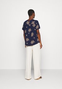 Blusa azul-marinho com padrão floral, mangas curtas e detalhe em gota nas costas, combinada com calças largas brancas leves.