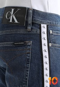 Tmavě modré džínové kalhoty s černou koženou nášivkou loga Calvin Klein Jeans na pase a bílým bočním pruhem se opakujícími se logy cK.