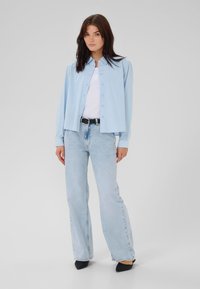 Camicia blu chiaro con bottoni, maniche lunghe, vestibilità rilassata; abbinata a jeans chiari a campana e tacchi neri a punta.