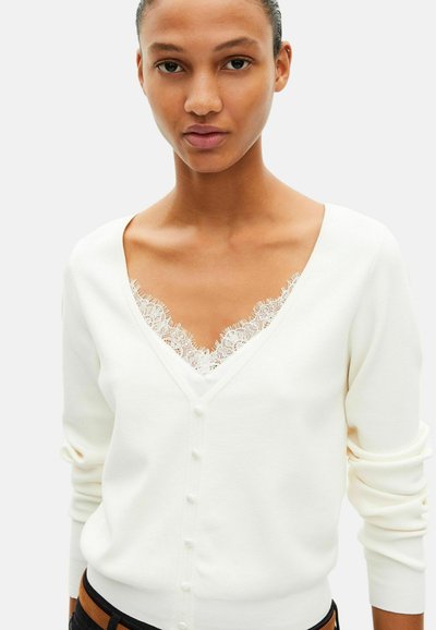 Hvid cardigan med V-udskæring og blonderkant ved halsen, med knaplukning foran og lange ærmer. Blød strikket stoftekstur.