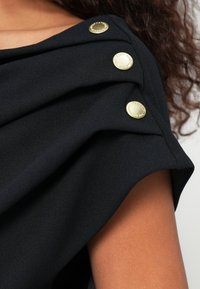 Μαύρο μπλουζάκι με draped ύφασμα στον ώμο, τρία χρυσά κουμπιά, ομαλή υφή και κοντά μανίκια.