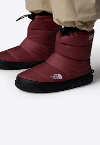 Bottes matelassées rouges avec une semelle en caoutchouc noire. Elles présentent un bout arrondi, un fermoir à cordon au niveau de la cheville et un logo sur le côté.