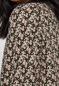 Blommigt mönster på tyg i mörkgrön, med små vita och rosa blommor. Slät textur med en lös passform, med fokus på axeldetaljer.