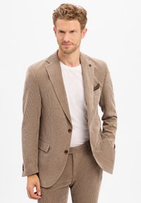 Man with beard and curly hair wearing a beige corduroy suit and white shirt, posing with one hand in pocket on white background.