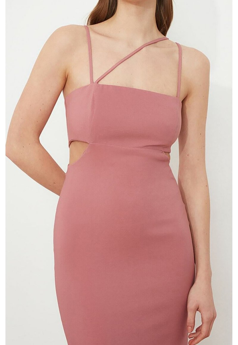 Femme portant une robe cintrée rose poussière avec des bretelles fines et un détail découpé sur le côté, sur un fond clair uni.