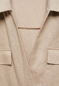 Prenda de lino beige con escote en V, que cuenta con dos bolsillos de parche y detalles de costura visibles. Tejido texturizado con un acabado mate.