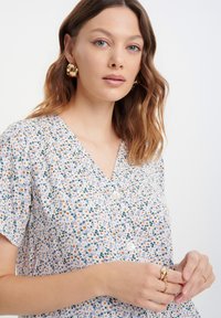 Vestido de estampado floral en púrpura claro con mangas cortas, escote en forma de V y botones blancos. Los accesorios incluyen pendientes de aro dorados y varios anillos.