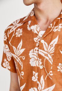 Camisa de lino de manga corta y cuello en color marrón óxido con patrones florales blancos. Cuenta con un bolsillo en el pecho y botones blancos.