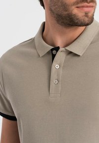 Béžové polo tričko s texturovanou tkaninou, třemi knoflíky a kontrastním černým lemem kolem límce. Detailní pohled zdůrazňuje límec a detaily šití.