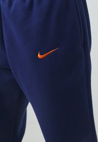 Námořnické modré tepláky vyrobené z měkké tkaniny; na levé nohavici je vyšitá oranžová ikona Nike; elastický pas pro pohodlí.